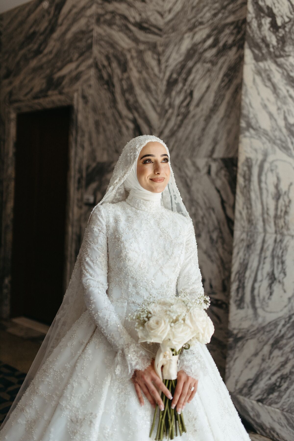 A bride in a high-necked beaded gown with a matching beaded hijab, holding white roses.