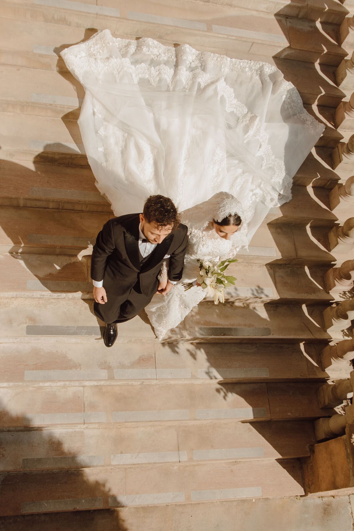 A bride and groom photographed from above on stone steps, her gown and train fanned out behind her.