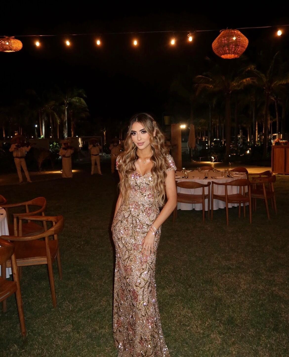 A woman in a full-length sequinned gown at an outdoor evening reception strung with lanterns.