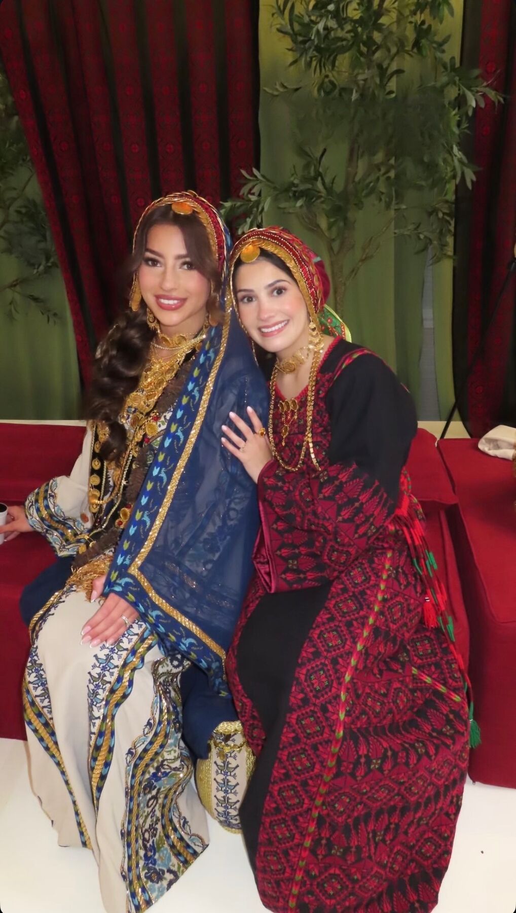 Two women seated together in traditional embroidered thobes with gold jewellery and headscarves.