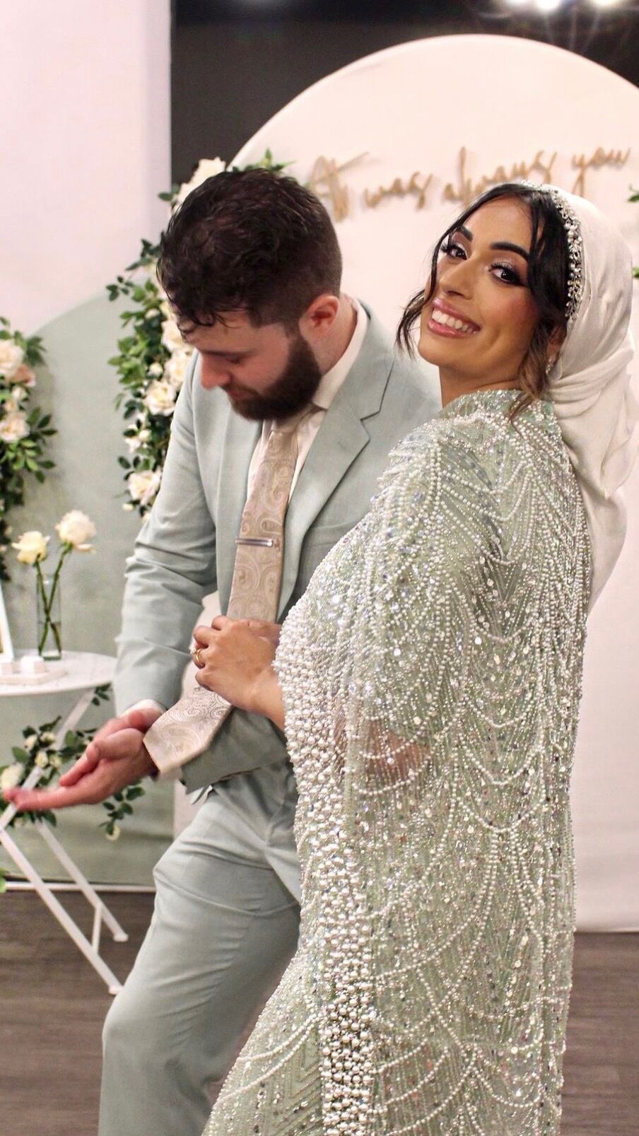 A bride in a pearl-beaded cape over her gown, turning to the camera beside the groom in a sage suit.