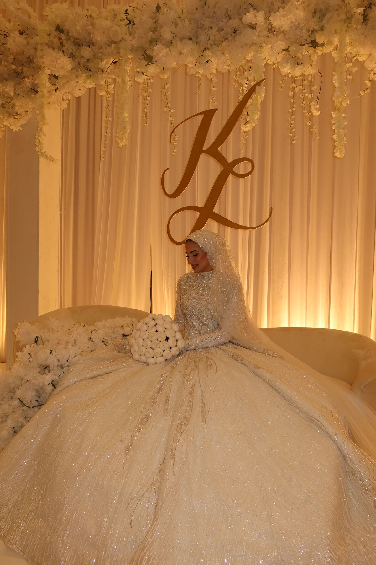 A bride in a sparkling full-skirted ball gown and matching beaded hijab, seated on a flower-framed stage beneath a gold monogram.