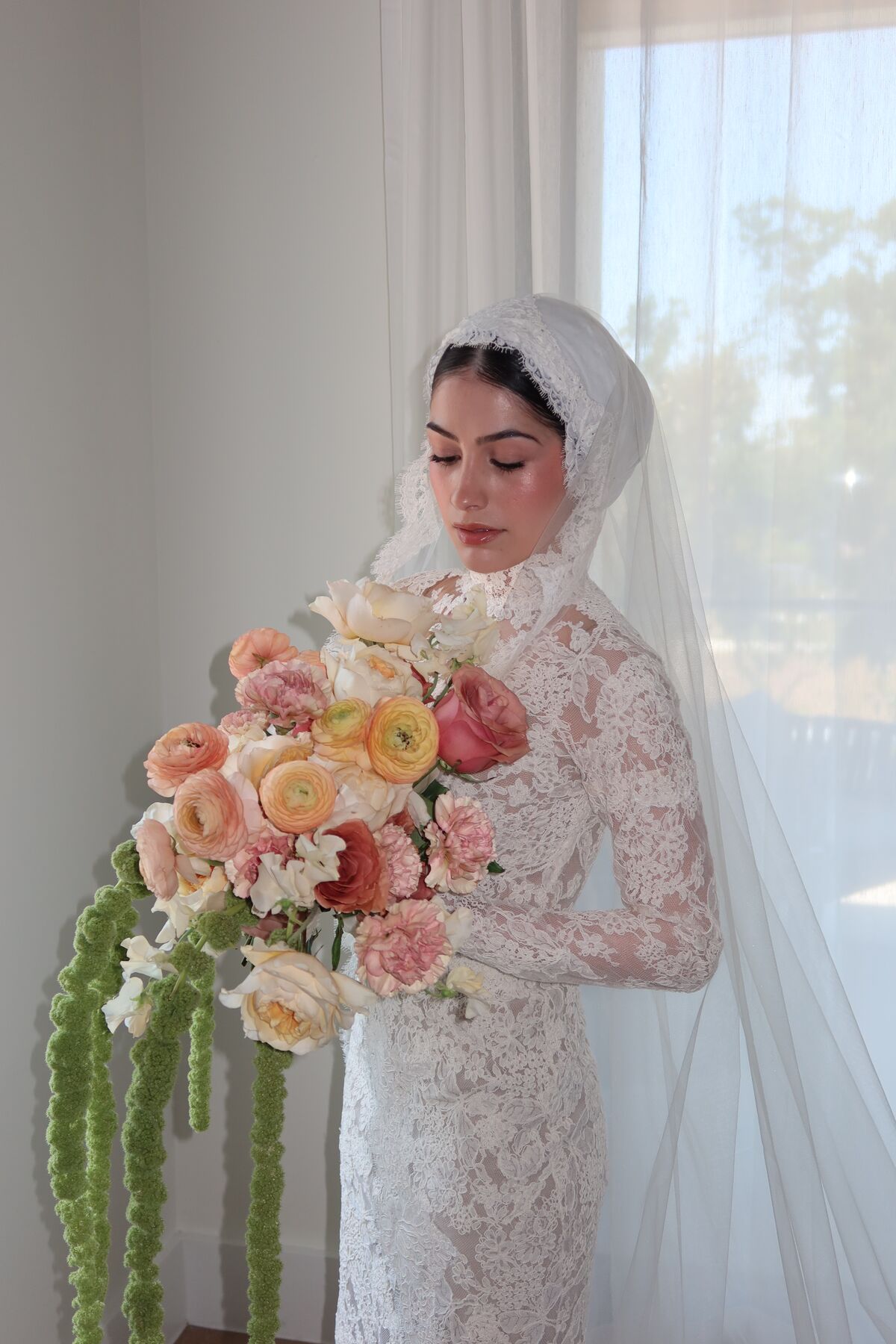A bride in a lace turban and long veil, holding a bouquet of peach and cream flowers.