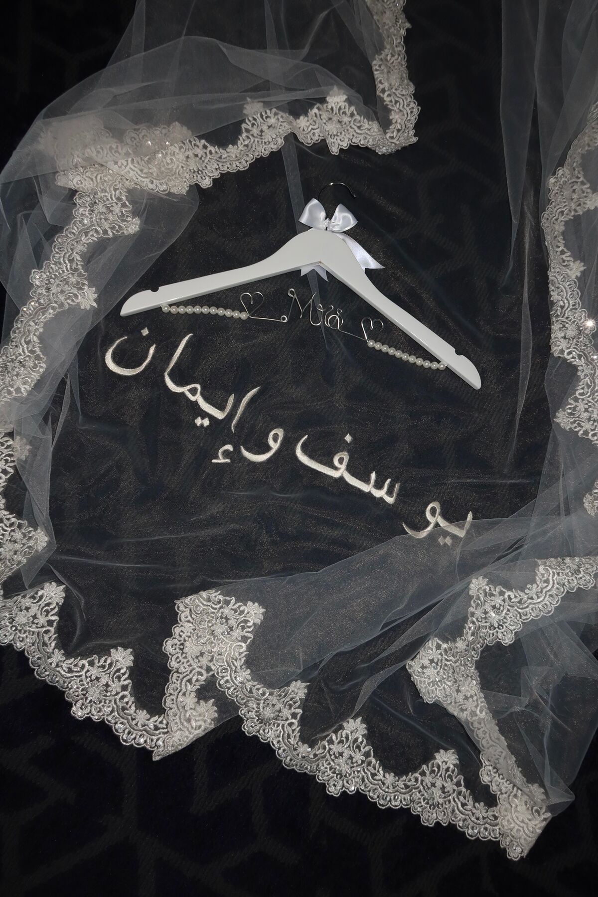 A veil on a white hanger against a dark ground, lace-edged and embroidered with two names in Arabic.