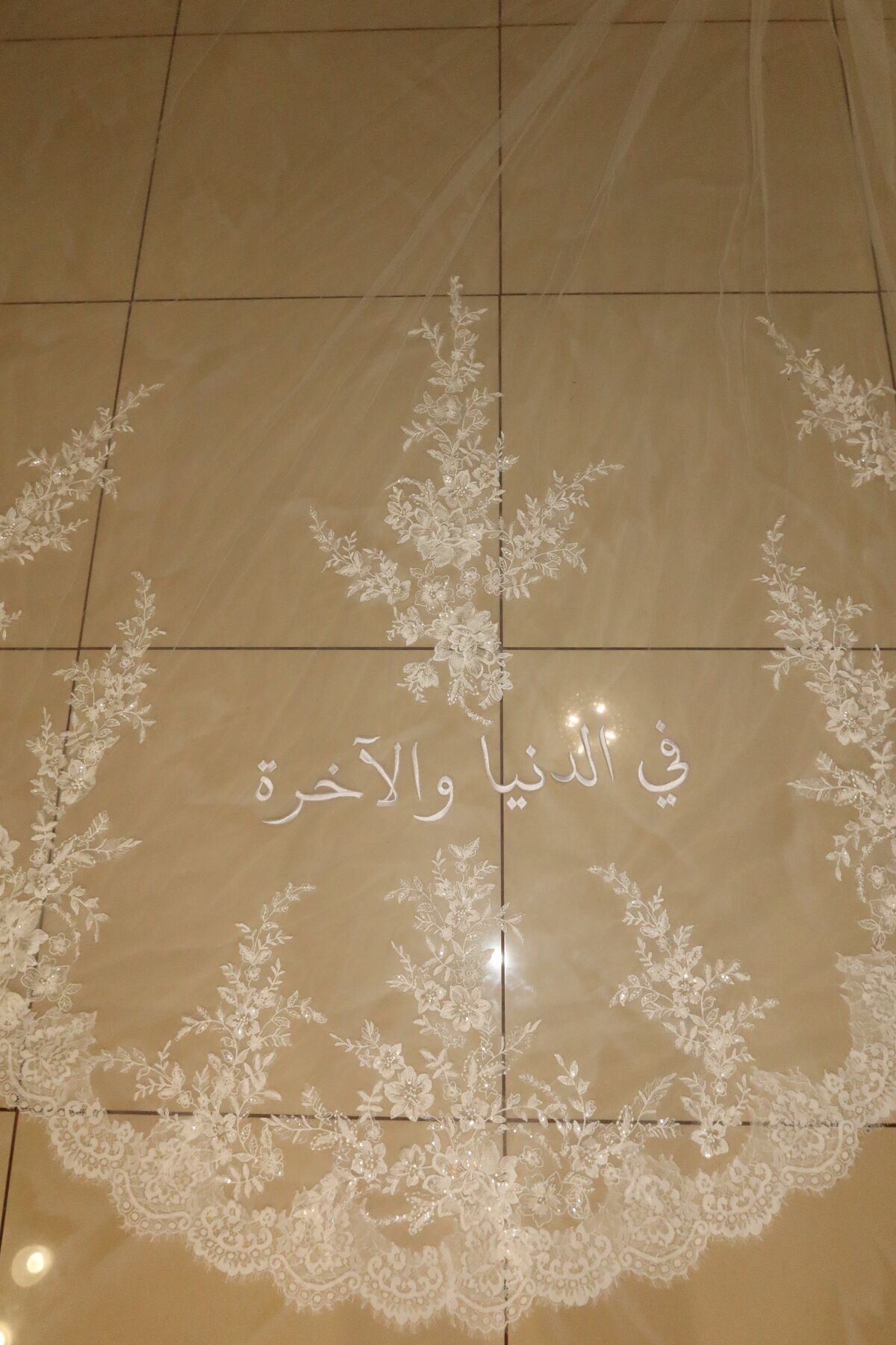 A long veil laid flat, its scalloped lace border framing an Arabic phrase embroidered across the tulle.